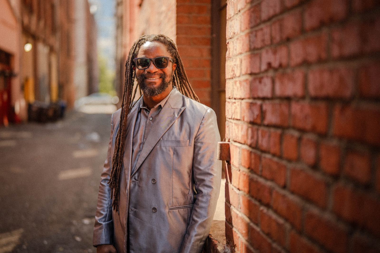 Simon Javan Okelo smiling in a silver suit, in a brick-walled alley