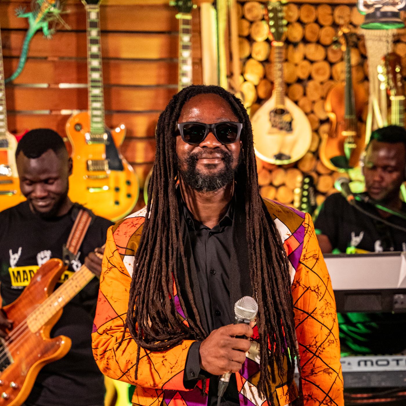 Simon Javan Okelo smiling with a microphone, his band and a wall of guitars behind him